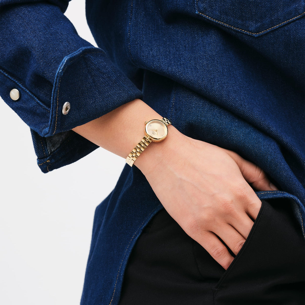 The gold Elan Lumine Unitone Gold women's watch with a round case and delicate 3-link bracelet is worn on the wrist with a dark blue denim blouse.