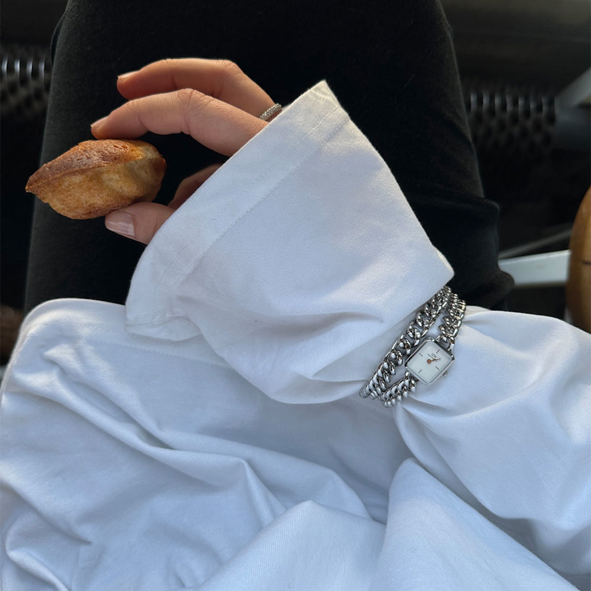 A person is wearing the Jolie & Chunky Chain Silver Set with a silver link bracelet watch and matching bracelet on their wrist over a white blouse, holding a pastry in their hand.