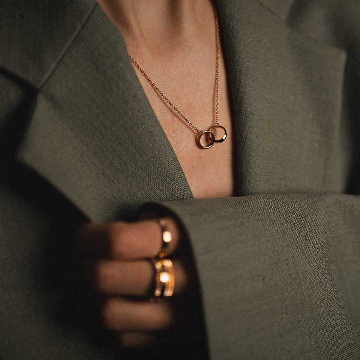 The Elan Unity necklace in rose gold with two interlinked rings is stylishly worn with a grey blazer and matching rings.