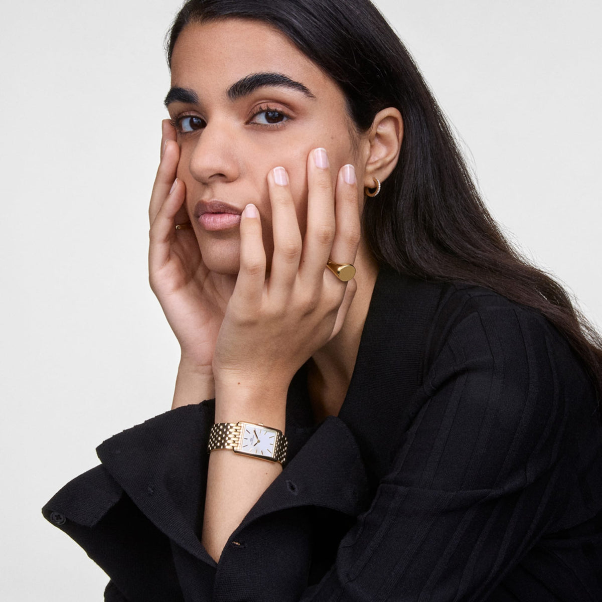 Woman with long dark hair wearing an elegant gold signet ring on her finger and a gold-coloured wristwatch with a black blazer.