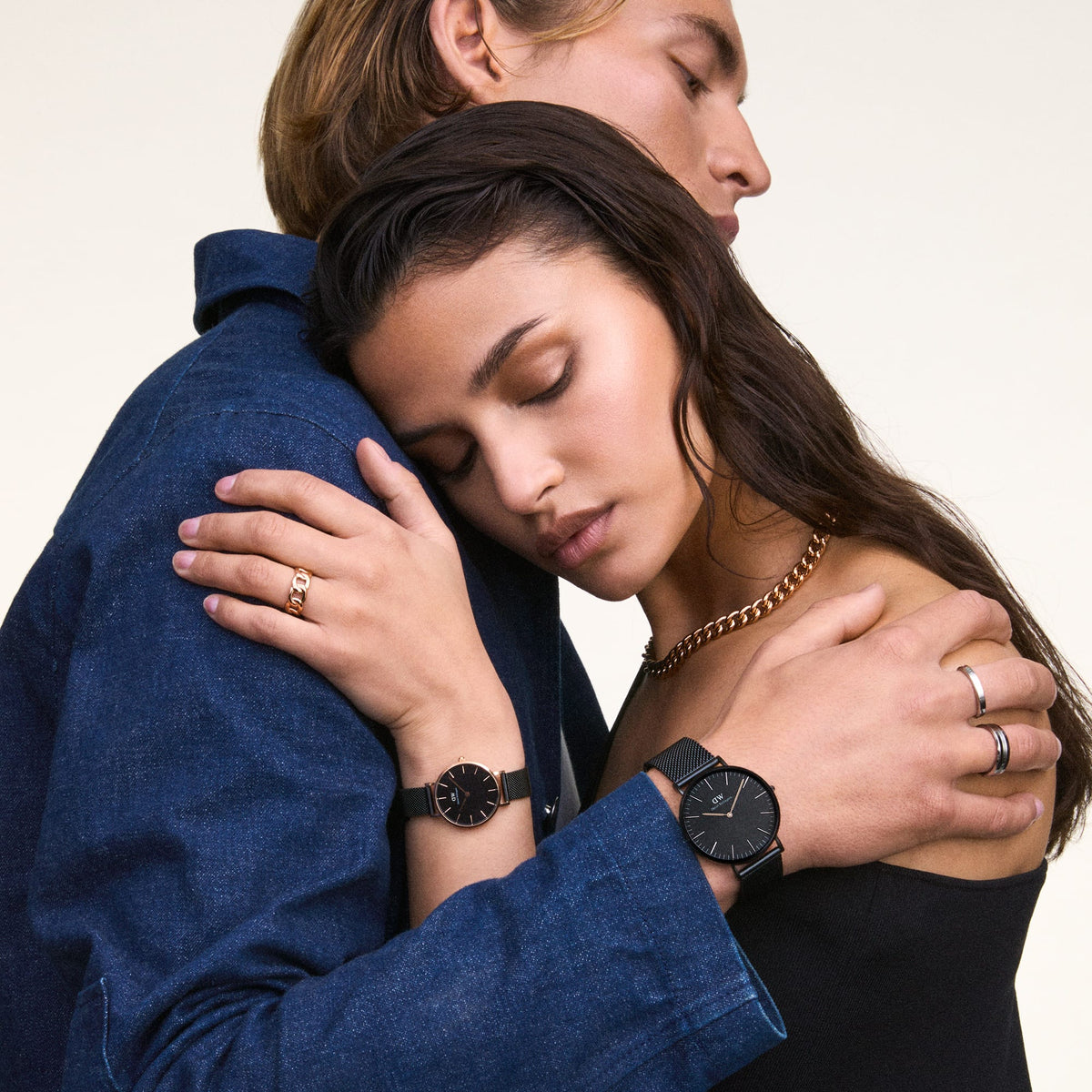 A woman wears the elegant Daniel Wellington Petite Ashfield with a black mesh strap and rose gold case on her wrist, while being closely embraced by a man wearing a matching watch.