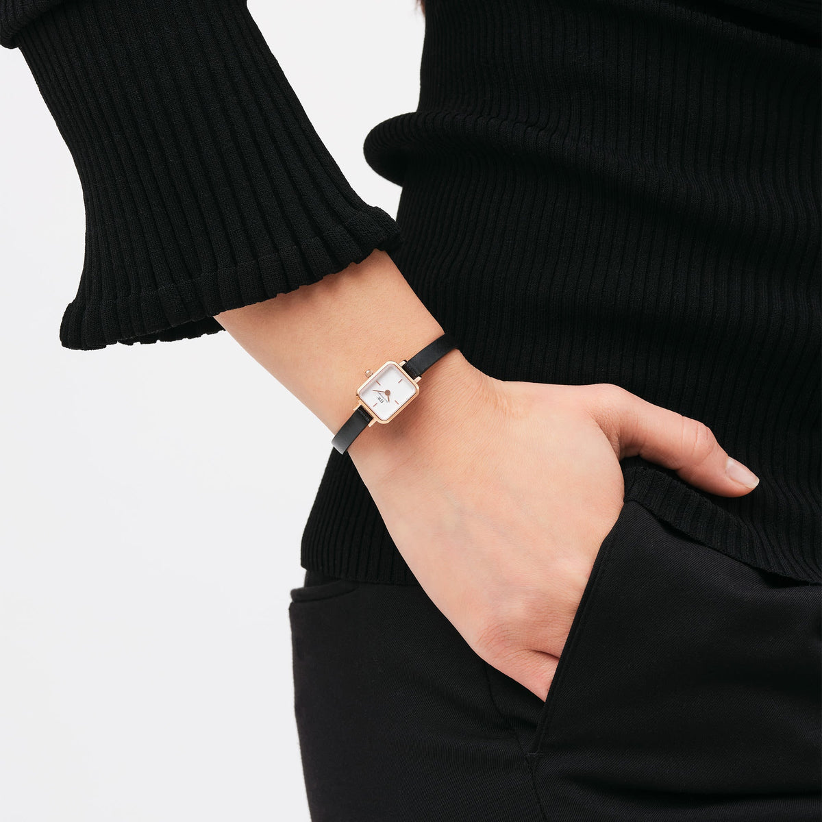 The Quadro Mini Sheffield Rose Gold women's watch with a rectangular rose gold case and black leather strap is worn on the wrist with a black outfit.