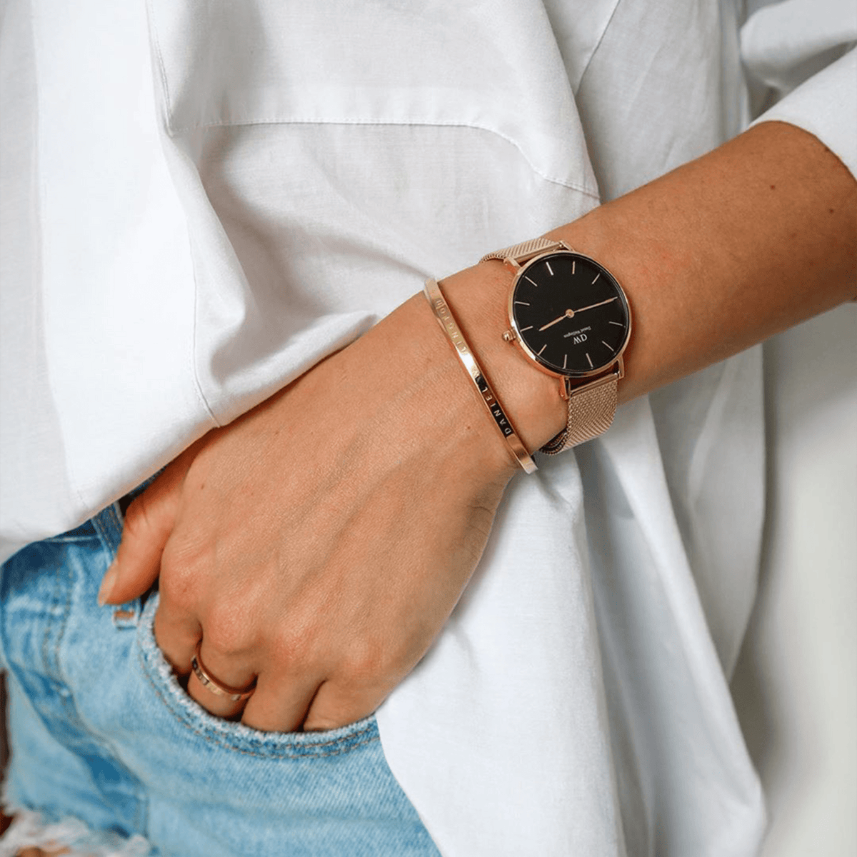 A woman wears the Daniel Wellington Petite Evergold Black with a black dial and gold-coloured mesh strap on her wrist, paired with a simple gold bangle and a casual outfit consisting of a white blouse and jeans.