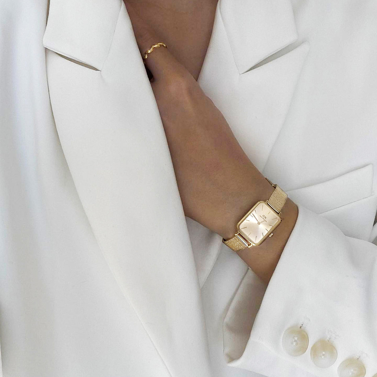 Elegant Quadro Pressed Unitone Gold ladies' watch with a rectangular case and gold-coloured mesh strap, worn with a white blazer.