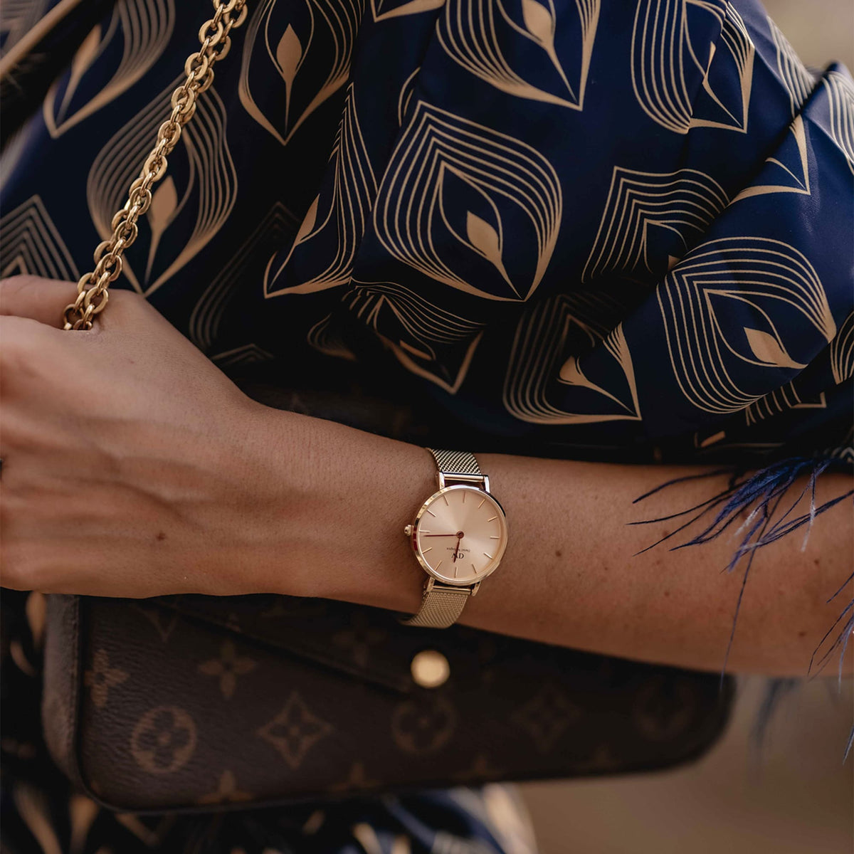 A woman wears the elegant Petite Unitone women's watch in rose gold with a fine mesh strap on her wrist, paired with a dark blue blouse and a designer handbag.