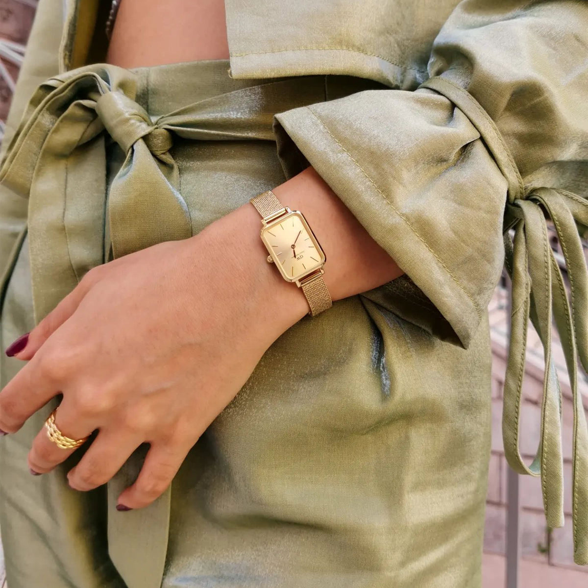 The Quadro Pressed Unitone Gold ladies' watch with a rectangular case and gold-coloured mesh strap is worn with an elegant green outfit.