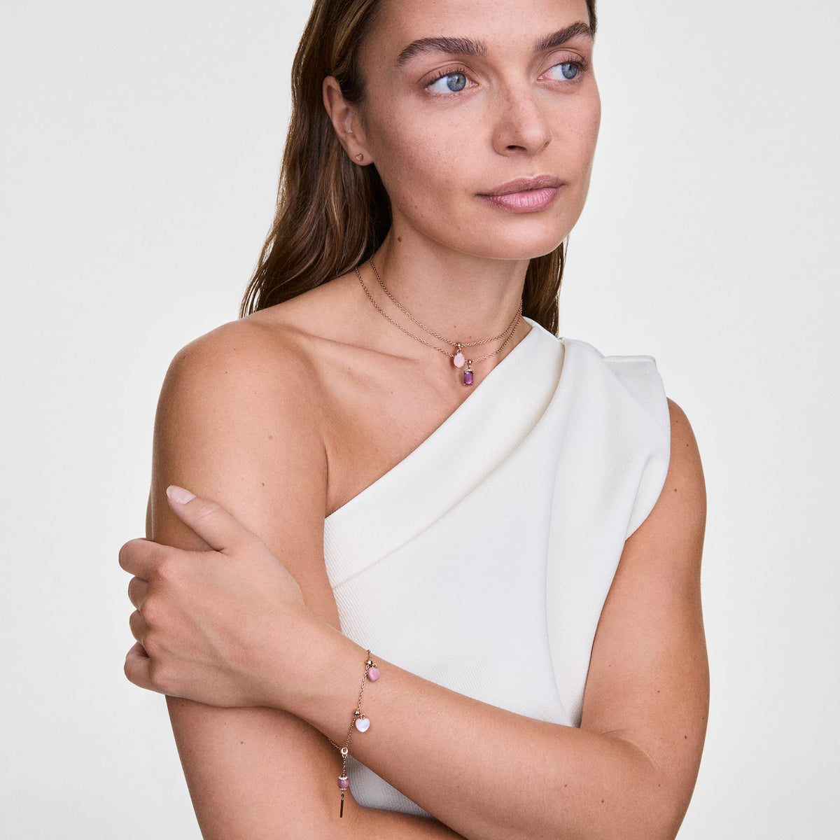 Mujer con vestido blanco luciendo el charm Rhodonite Heart Gold Charm en un collar y una pulsera dorados con piedras de rodonita rosa