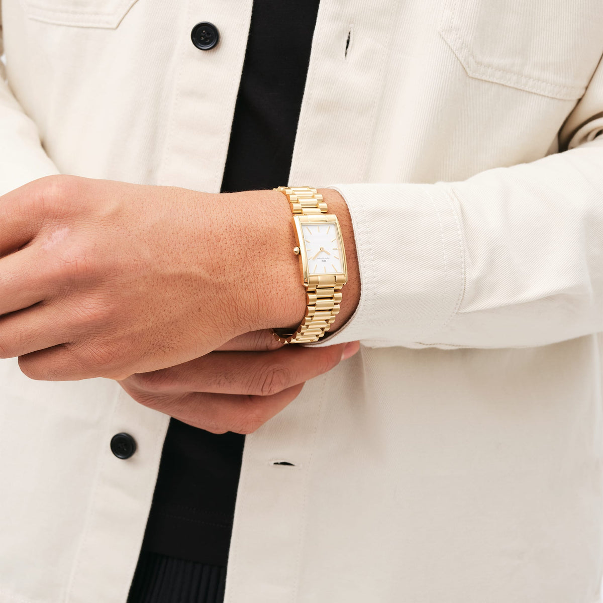 A gold Daniel Wellington Bound 3-Link ladies' watch with a rectangular case and white dial is worn on the wrist with a cream-coloured shirt.