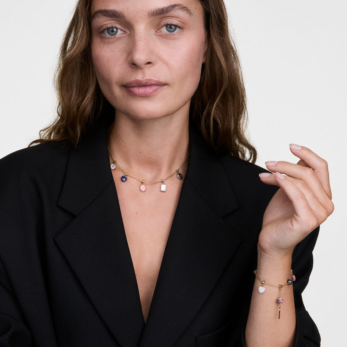 La modella indossa una collana e un bracciale dorati con charm componibili, tra cui il ciondolo *Blue Spot Oval Gold Charm* con pietra naturale blu, abbinati a una giacca nera su sfondo neutro.