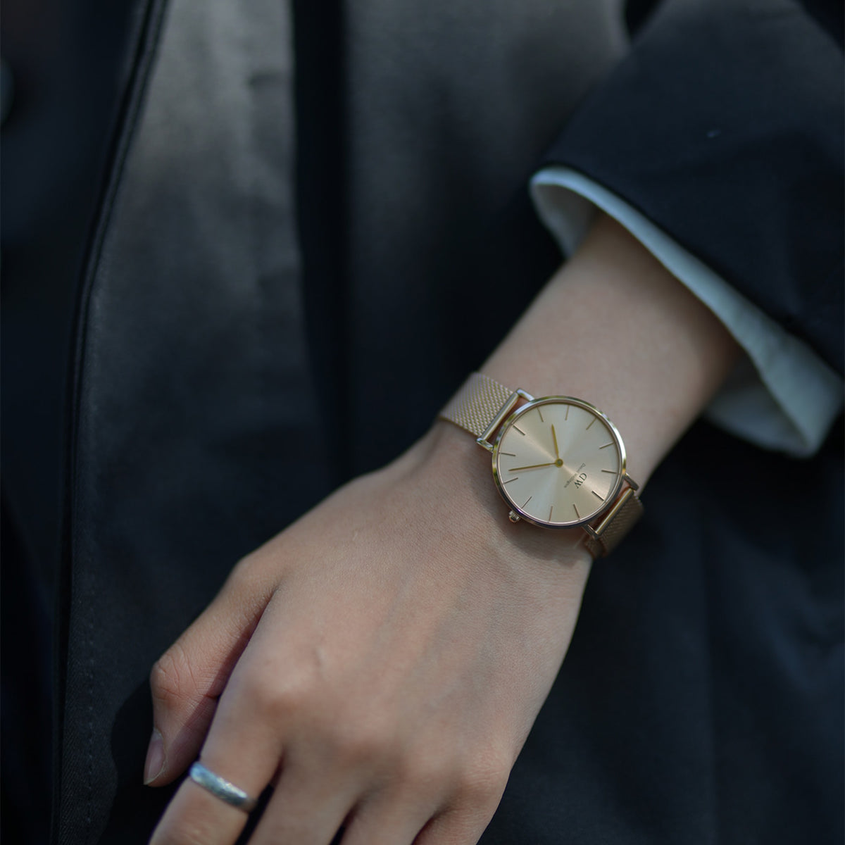 The gold Daniel Wellington Petite Unitone women's watch with a fine mesh strap is worn elegantly on the wrist with a dark, classic outfit.
