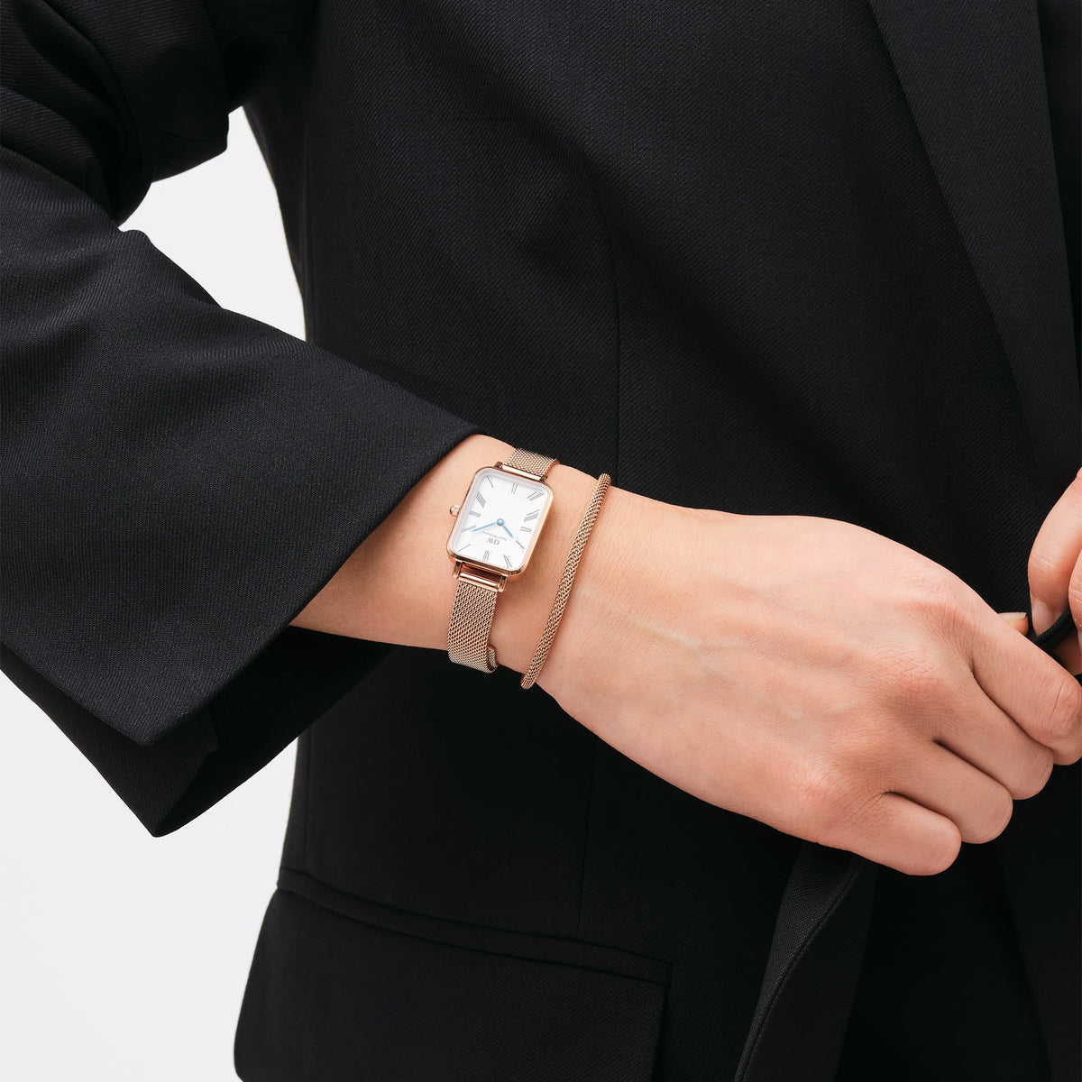 Elegant ladies' watch Quadro Roman Numerals Melrose with a rectangular rose gold case and white dial on the wrist, paired with a matching bracelet and black blazer.