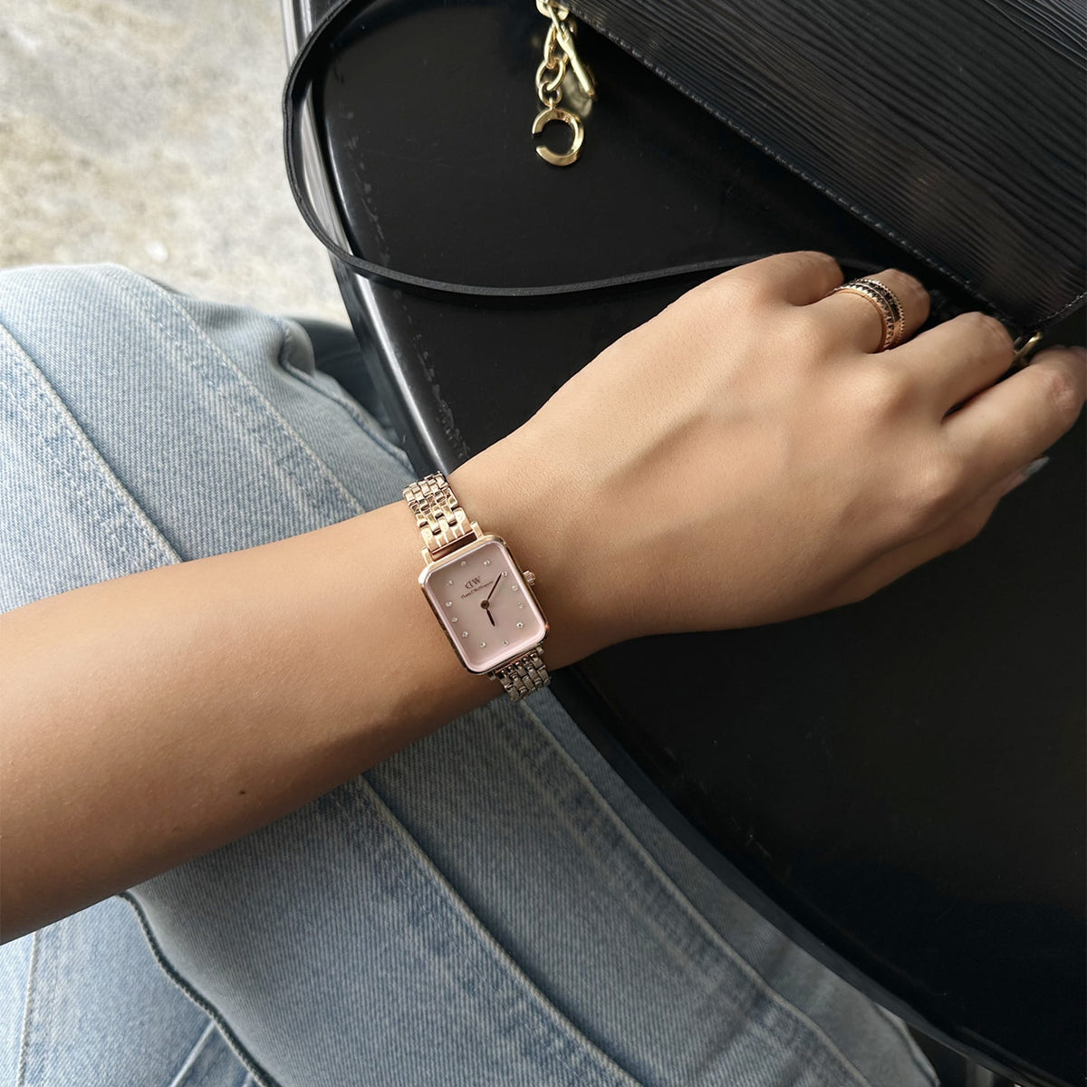 Orologio Quadro Lumine da polso, abbinato a una borsa nera e anelli in oro, che mette in risalto il quadrante rosa impreziosito da cristalli e il cinturino a cinque maglie in oro rosa su un paio di jeans chiari.