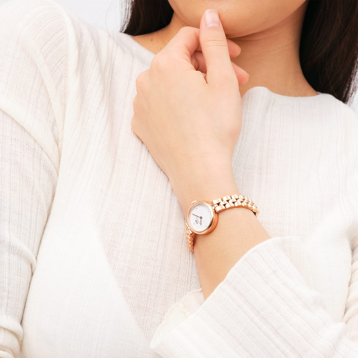 The Elan Lumine Rose Gold women's watch with a round case, white dial and crystal indices is worn on a slender wrist with a cream-coloured top.