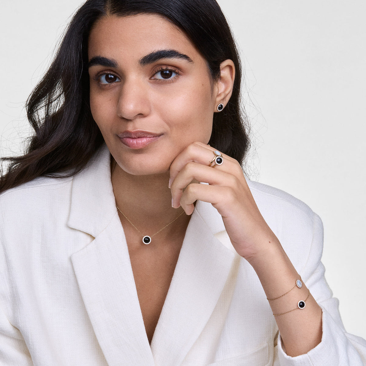 A woman wears the Audrey Necklace Black Agate Gold with a round pendant made of black agate and white zirconia crystals, paired with matching jewellery and an elegant white blazer.
