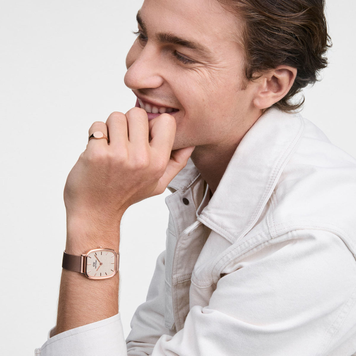 A man wears a classic rose gold signet ring with a polished finish on his finger, stylishly pairing it with a matching watch and a light-coloured jacket.