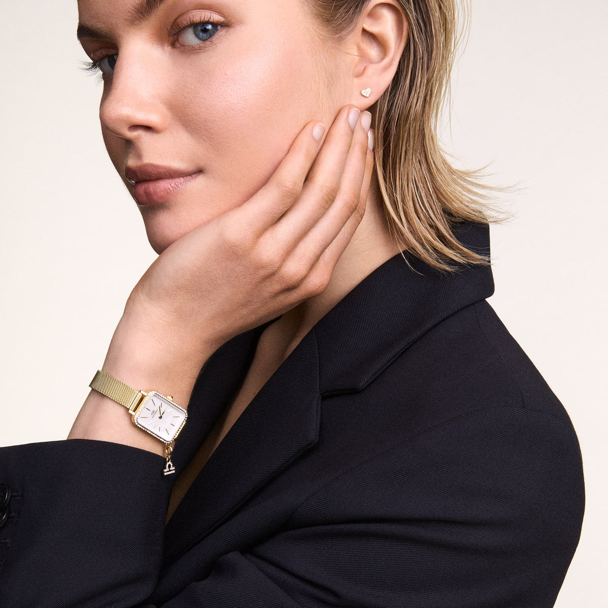 A woman elegantly wears the Quadro Crystal Zodiac Evergold women's watch with a rectangular case, sparkling crystals, white mother-of-pearl dial, and gold-coloured mesh strap on her wrist with a black blazer.