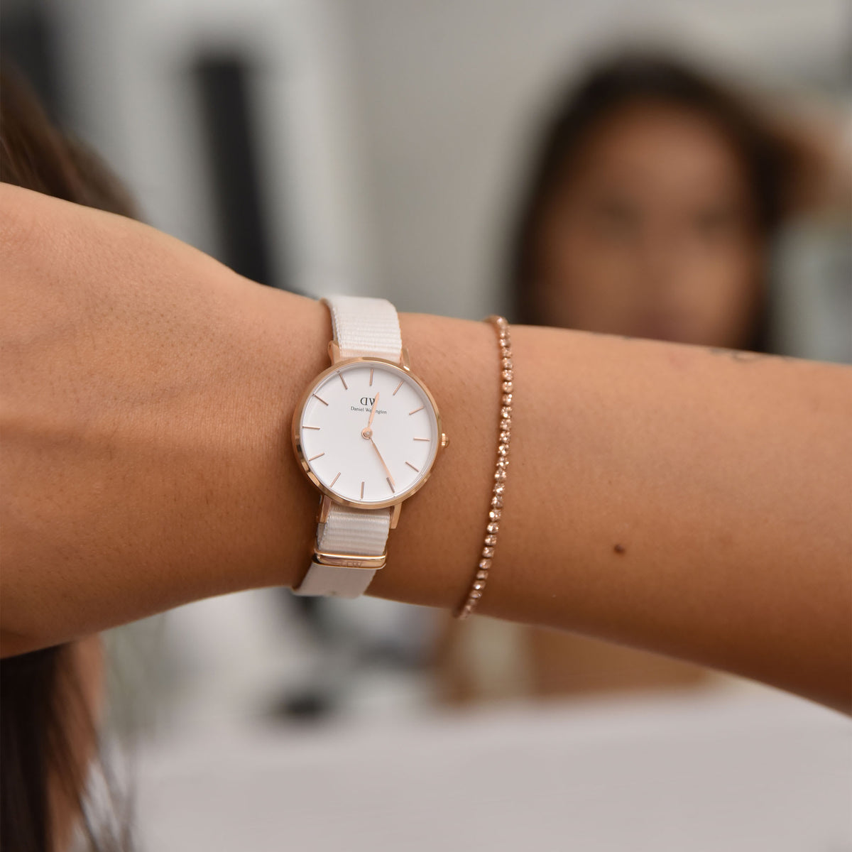 Bracciale tennis con cristalli color pesca indossato al polso accanto a un orologio Daniel Wellington dal cinturino bianco e cassa in oro rosa.