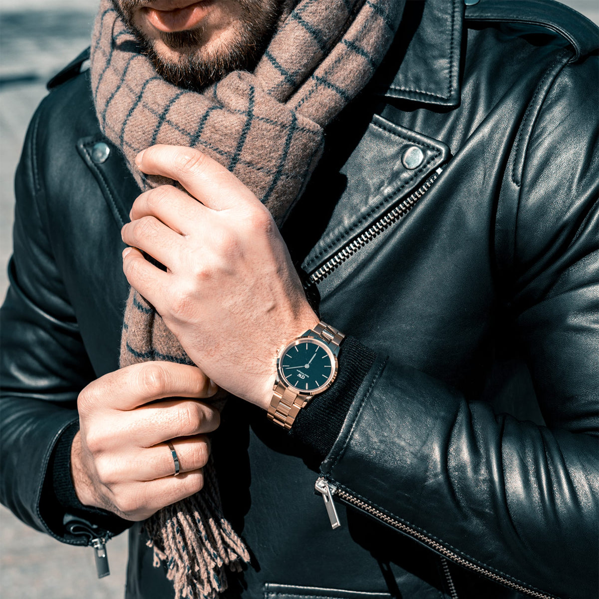 The Daniel Wellington Iconic Link watch in rose gold with a black dial is worn on a wrist with a black leather jacket and a checked scarf.
