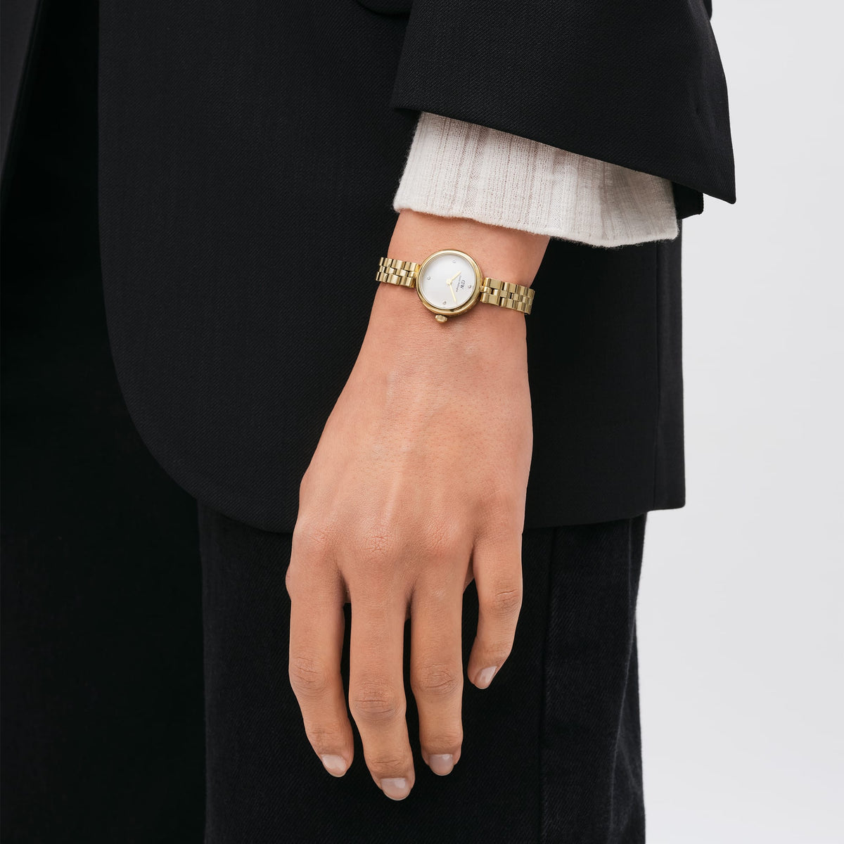 The Elan Lumine Gold women's watch with a gold-coloured stainless steel bracelet and white dial with crystal indices is worn elegantly on the wrist with a black blazer.