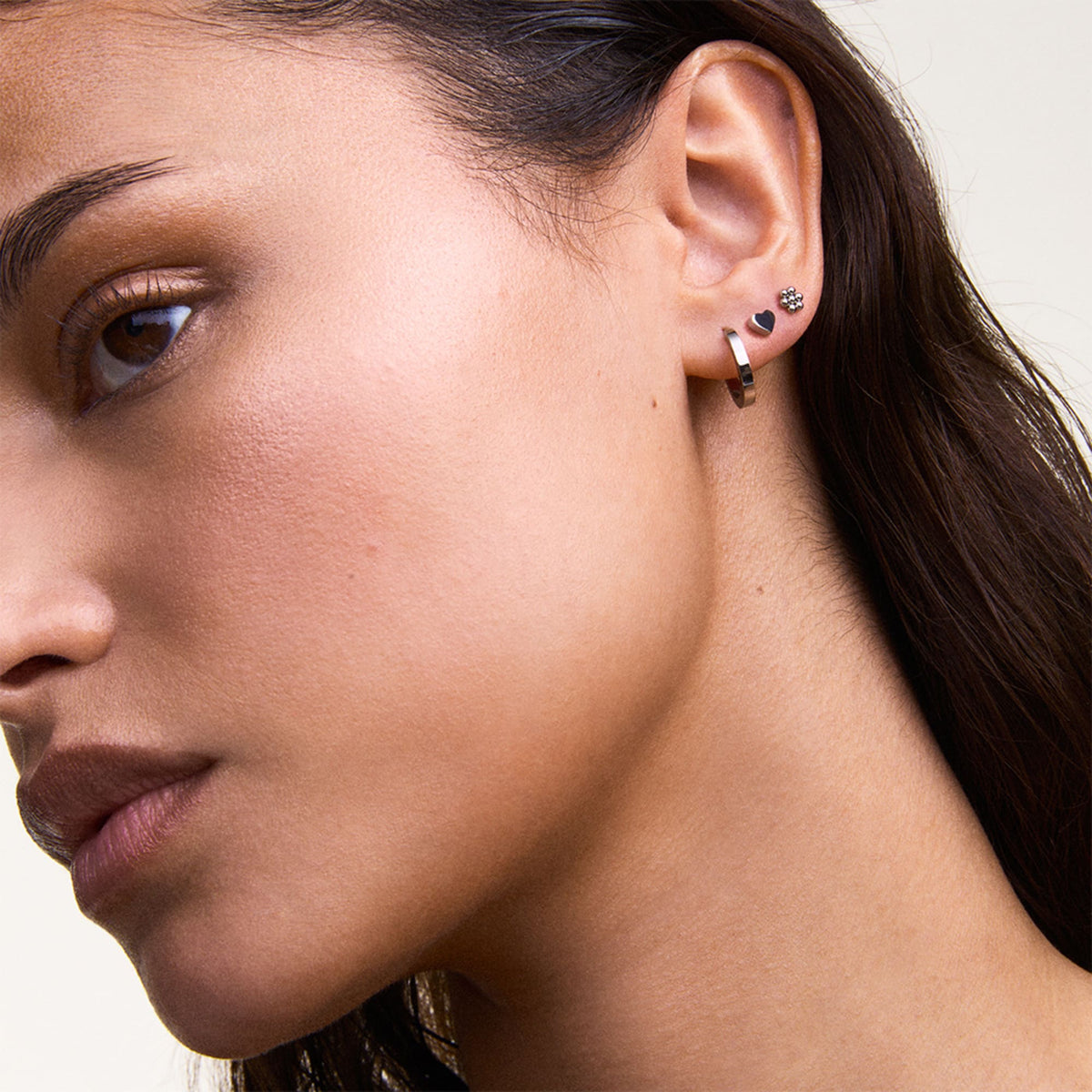 Mujer luciendo el set de pendientes de plata con aro minimalista en el lóbulo y pendientes de botón con forma de corazón y flor en las perforaciones superiores