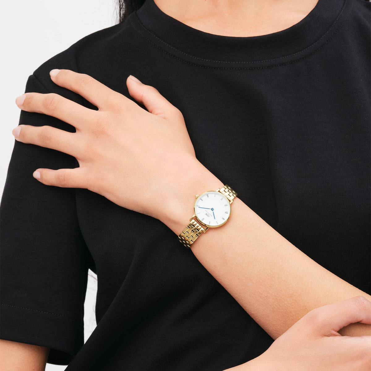 A delicate gold women's watch with a 5-link stainless steel bracelet, white dial, Roman numerals and blue hands is worn on the wrist of a woman wearing a black T-shirt.