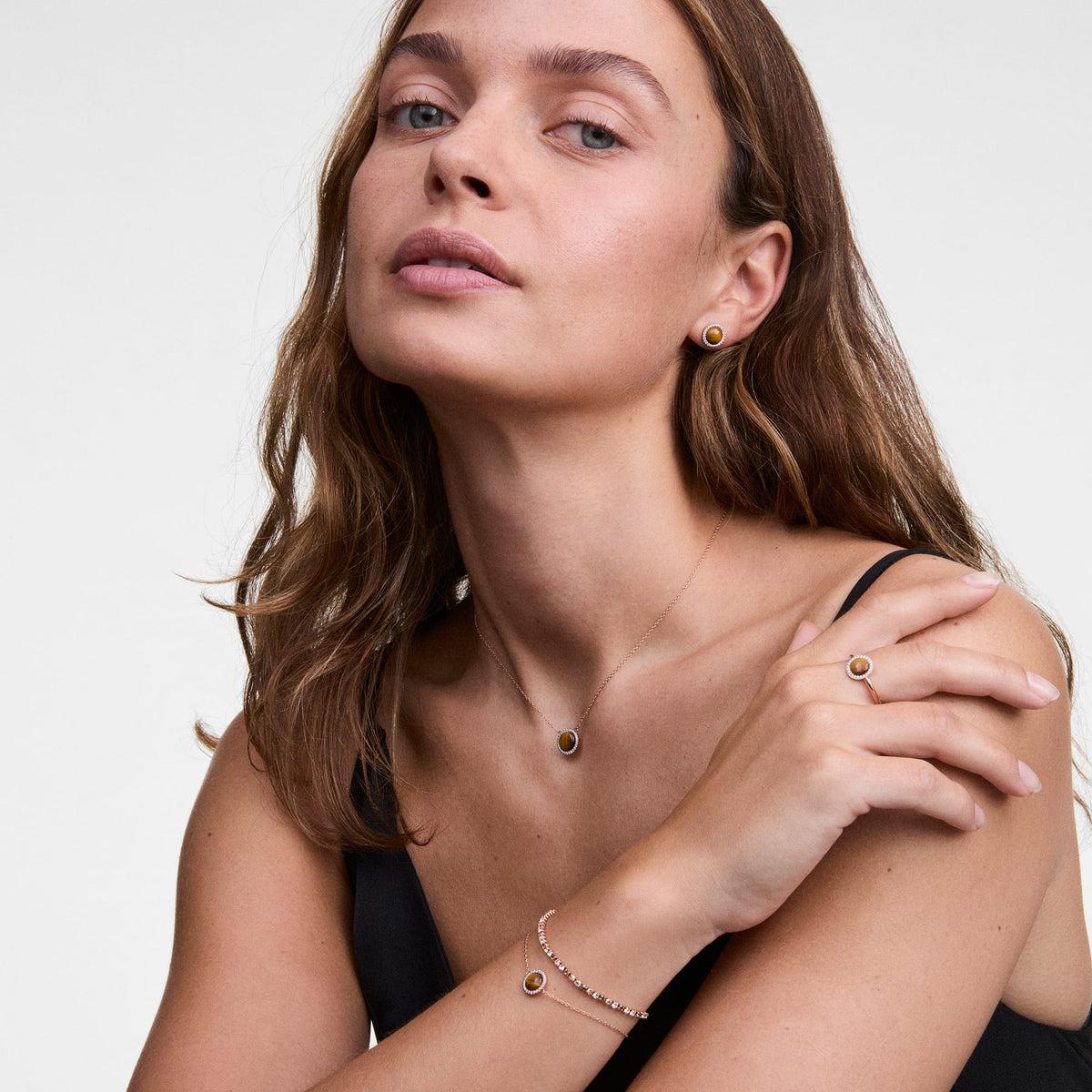 A woman wears Audrey stud earrings with tiger's eye in silver, paired with a matching necklace, bracelet and ring, all elegantly harmonised by the brown gemstone and sparkling accents.