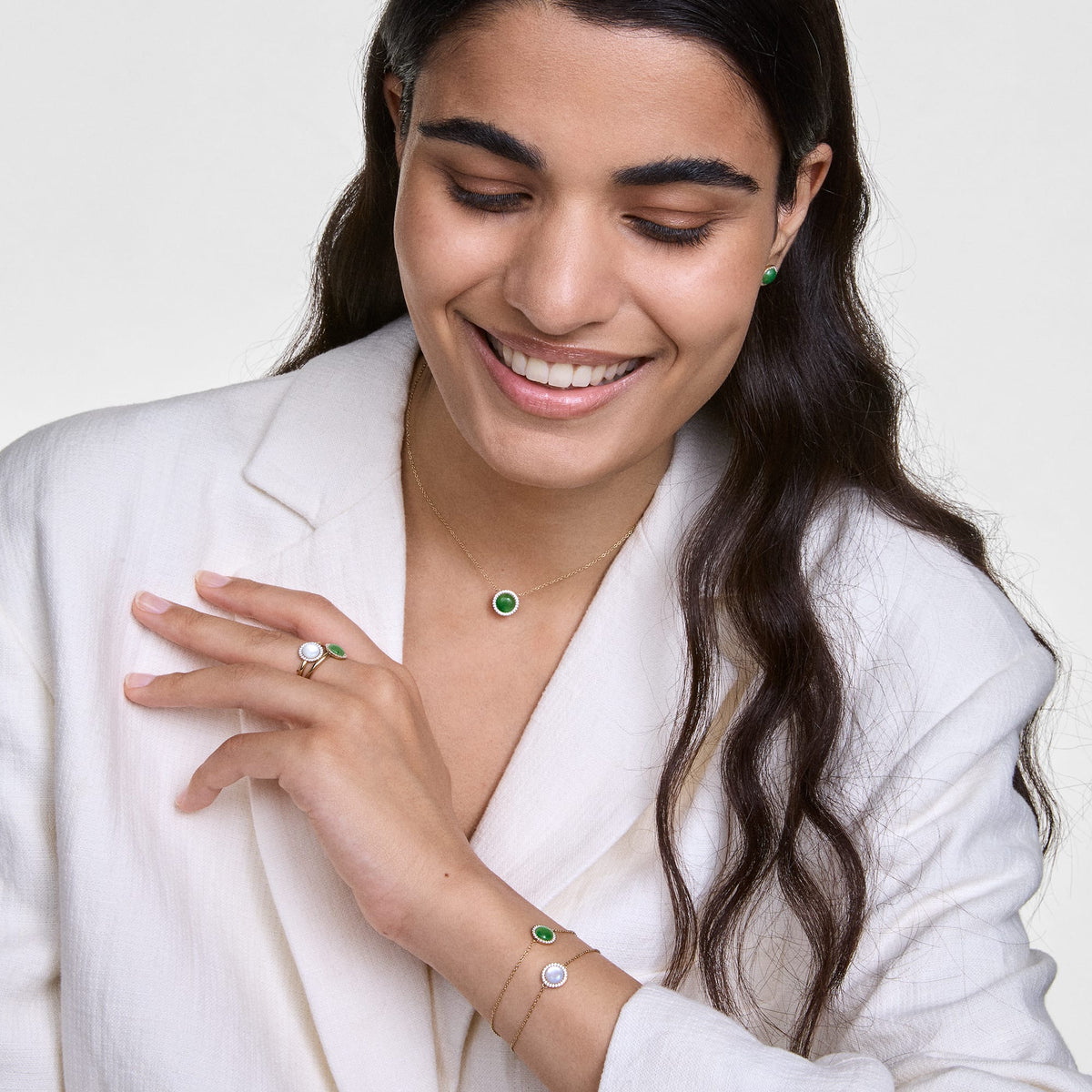 A woman wears an elegant gold-coloured bracelet with a round mother-of-pearl pendant and sparkling crystals on her wrist, combined with other jewellery pieces and a white blazer.