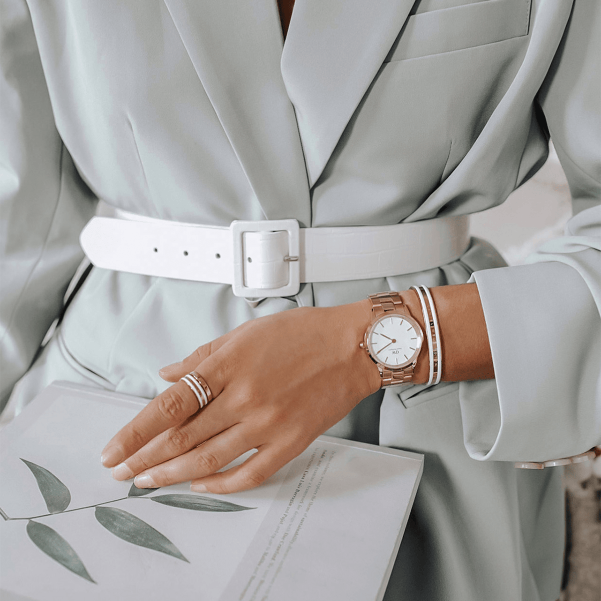 The Daniel Wellington Iconic Link women's watch in rose gold with a white dial is stylishly worn on the wrist with an elegant light-coloured blazer and matching accessories.