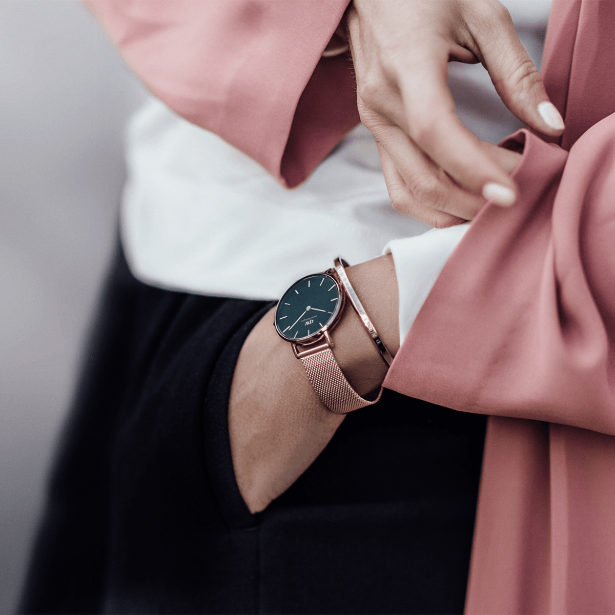 The Daniel Wellington Petite Melrose women's watch with black dial and rose gold mesh strap is stylishly worn on the wrist with a pink blazer and bangle.