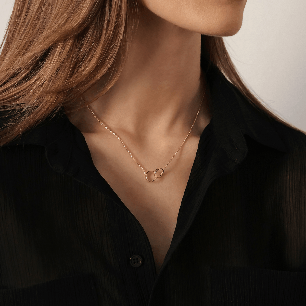 The Elan Unity necklace in rose gold with two delicately interwoven rings is worn elegantly around a woman's neck with a black blouse.