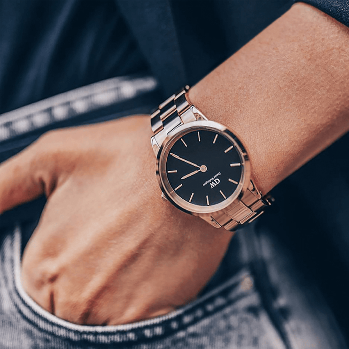The Daniel Wellington Iconic Link watch in rose gold with a black dial is casually worn on the wrist with jeans and dark clothing.