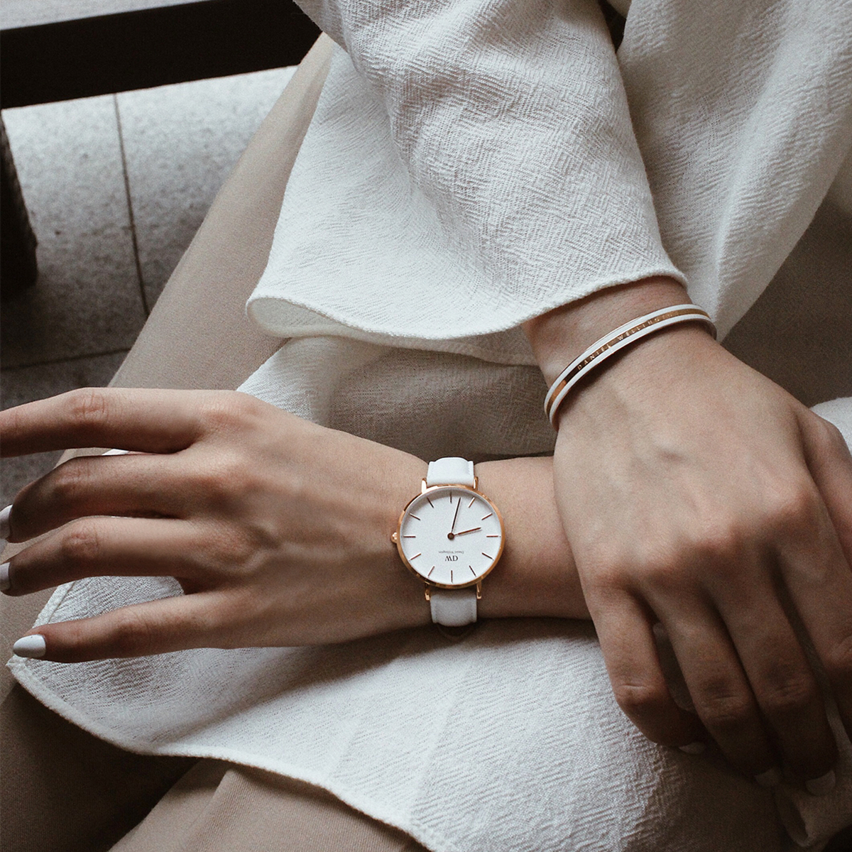 A woman wears the elegant Petite Bondi Rose Gold ladies' watch with a white leather strap and rose gold case on her wrist, paired with a matching bangle for a stylish, light-coloured outfit.
