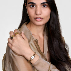 A woman wears the elegant Jolie Chain Gold women's watch with a gold-coloured chain bracelet and a rectangular white dial on her wrist, paired with gold rings and a sheer blouse.