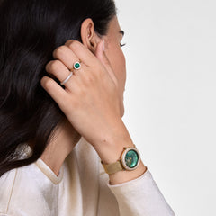 A woman wears the Audrey Ring Malachite Gold with a round green malachite and zirconia crystals on her finger, paired with a gold-coloured watch and another ring for an elegant, modern look.