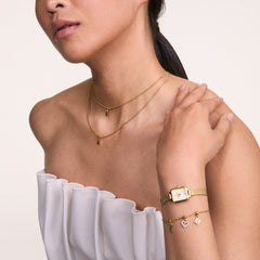 A woman wears the Quadro Mini Evergold Champagne ladies' watch with a rectangular gold-coloured case and mesh strap on her wrist, paired with delicate gold jewellery and an elegant white top.