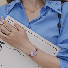 A woman wears the Petite Lumine 5-Link Melrose ladies' watch in rose gold with a pink mother-of-pearl dial and crystal indices on her wrist, paired with a blue blouse and subtle jewellery.