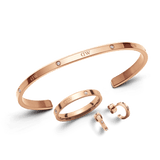Set di gioielli **Lumine Jewelry Set** in acciaio inossidabile con placcatura in oro rosa, composto da un bracciale rigido aperto, un anello sottile e orecchini a cerchio semicircolari. Tutti i pezzi sono impreziositi da cristalli incastonati e presentano il logo inciso.