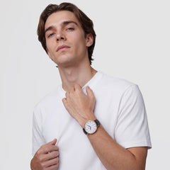Un uomo indossa un orologio con cassa dorata, quadrante bianco arricchito da numeri romani e cinturino in pelle marrone scuro effetto coccodrillo. L'accessorio è abbinato a una t-shirt bianca su uno sfondo neutro.
